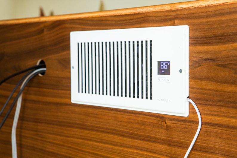 Puede incluir: Una rejilla de ventilaci&oacute;n AC Infinity blanca con lamas verticales y una pantalla digital que muestra 30&deg;C. La rejilla est&aacute; montada sobre una superficie de madera oscura. Un cable blanco sale de la rejilla.