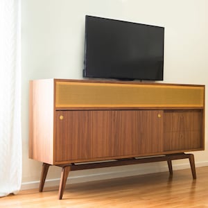 Puede incluir: Un mueble de TV moderno de mediados de siglo con acabado en madera oscura, panel de altavoz de malla dorada y herrajes de lat&oacute;n. Encima hay un televisor de pantalla plana negro. El mueble est&aacute; sostenido por patas anguladas.