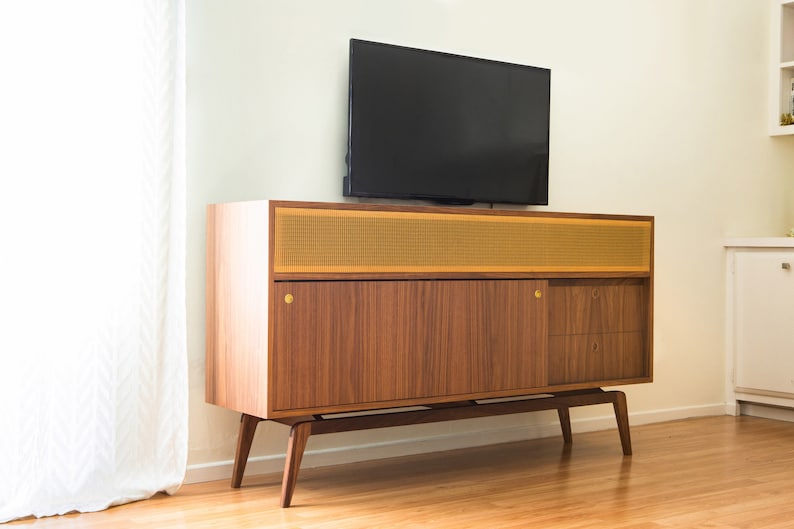 Puede incluir: Un mueble de TV moderno de mediados de siglo con acabado en madera oscura, panel de altavoz de malla dorada y herrajes de lat&oacute;n. Encima hay un televisor de pantalla plana negro. El mueble est&aacute; sostenido por patas anguladas.