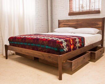 Könnte beinhalten: Ein Holzbettgestell mit einem Kopfteil und einem Fußteil aus Walnussholz. Das Bett hat eine rote, schwarze und türkisfarbene gemusterte Decke auf der weißen Bettwäsche. Das Bettgestell hat zwei Schubladen unten.
