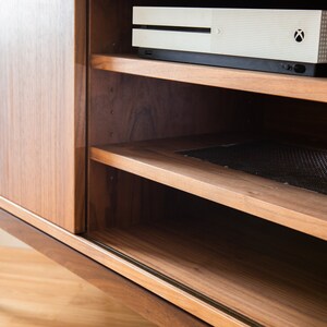 Puede incluir: Un mueble de entretenimiento de madera de estilo moderno de mediados de siglo con una puerta corredera y estanter&iacute;as abiertas. La unidad est&aacute; hecha de madera de tonos c&aacute;lidos y presenta una consola en la parte superior. Los estantes est&aacute;n dise&ntilde;ados para contener componentes multimedia.