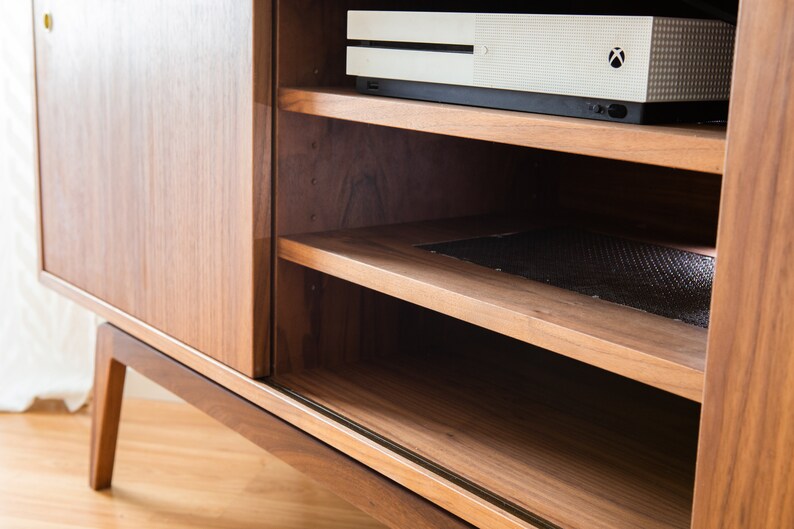 Puede incluir: Un mueble de entretenimiento de madera de estilo moderno de mediados de siglo con una puerta corredera y estanter&iacute;as abiertas. La unidad est&aacute; hecha de madera de tonos c&aacute;lidos y presenta una consola en la parte superior. Los estantes est&aacute;n dise&ntilde;ados para contener componentes multimedia.