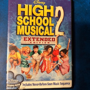 Może przedstawiać: Płyta DVD z filmu "High School Musical 2: Extended Edition" Disneya. Okładka przedstawia tytuł w kolorze żółtym i obsadę skaczącą do basenu. Obudowa DVD jest czarna z logo Disney Channel i tekstem "Includes Never-Before-Seen Music Sequence."