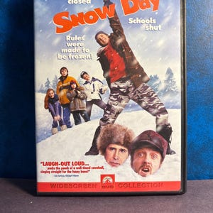 May include: A DVD case for the movie "Snow Day" with a winter theme. The cover features a group of children and adults in winter clothing, with the title in large orange letters. Text includes "Roads closed" and "Schools shut."