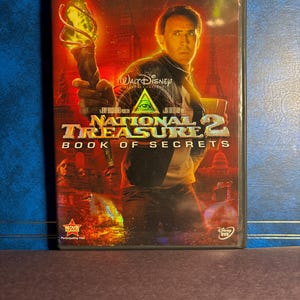 Könnte beinhalten: DVD des Films "Das Vermächtnis 2: Das Vermächtnis von Benjamin Gates" mit Nicolas Cage. Das Cover zeigt Cage mit einer Fackel vor einem Hintergrund von Stadtlandschaften, darunter der Eiffelturm und Big Ben. Das Walt Disney Logo ist ebenfalls zu sehen.