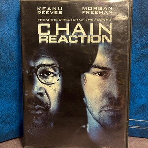 Może przedstawiać: Pudełko DVD filmu "Chain Reaction" z Keanu Reevesem i Morganem Freemanem. Tytuł jest dużymi, pogrubionymi białymi literami. Tło jest ciemne z twarzami aktorów.