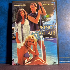 Może przedstawiać: Pudełko DVD filmu "Prince of Bel Air" z Markiem Harmonem i Kirstie Alley. Okładka przedstawia trzy osoby w strojach kąpielowych w pobliżu basenu. Tekst na pudełku brzmi "He has it all. Except the girl."