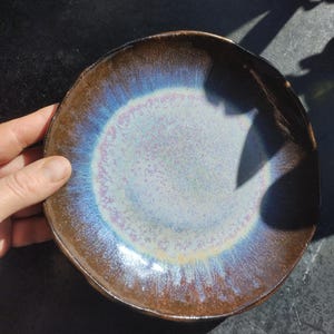 Może przedstawiać: Ręcznie robiony ceramiczny talerz z unikalnym iryzującym szkliwem. Talerz ma brązową krawędź, która przechodzi w niebiesko-fioletowy środek. Nieregularny kształt talerza dodaje mu artystycznego uroku. Idealny do serwowania potraw lub jako element ekspozycji.