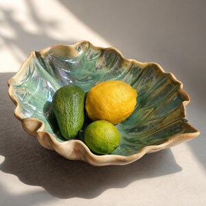 Può includere: Una ciotola in ceramica a forma di foglia con smalto turchese contiene un avocado, un limone e un lime. La ciotola ha una superficie strutturata e un bordo marrone chiaro. I frutti sono disposti all'interno della ciotola.