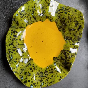 May include: A unique, free-form ceramic dish with a vibrant design. The dish features a bright yellow center surrounded by a rim of speckled green and black. Small blue dots add detail to the rim. The dish is an artistic piece.