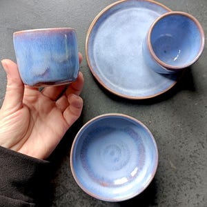 Puede incluir: Un juego de vajilla de cerámica que incluye dos tazas cilíndricas y un plato pequeño redondo, todo acabado con un esmalte azul matizado y bordes de color marrón rojizo. Las piezas presentan una forma lisa y redondeada con sutiles degradados.