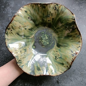 Può includere: Una ciotola in ceramica fatta a mano con una forma organica unica. La ciotola presenta una miscela di smalto verde, beige e nero, creando un effetto marmorizzato. Il bordo ha un bordo marrone scuro. La ciotola è tenuta da una mano.