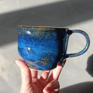 Può includere: Una tazza in ceramica fatta a mano con una glassa blu intenso. La tazza ha una forma arrotondata con un manico grande e un bordo marrone a contrasto. La tazza è tenuta in una mano, mostrando le sue dimensioni e il suo design.