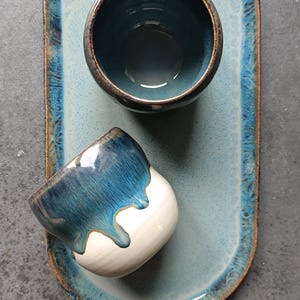Puede incluir: Una bandeja ovalada de cerámica con un centro azul claro moteado y un borde azul oscuro, acompañada de dos tazas a juego. Una taza está en posición vertical mostrando un interior azul profundo, mientras la otra descansa de lado con base blanca.