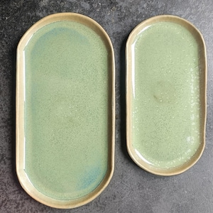 May include: Two oval-shaped ceramic trays with a light green speckled glaze and a smooth, tan-colored rim. The trays feature a glossy finish and a slightly irregular, organic shape, with one tray noticeably larger than the other.