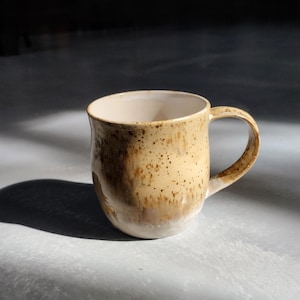 Pode incluir: Uma caneca de cerâmica marrom e branca com manchas, de forma arredondada e com uma alça.