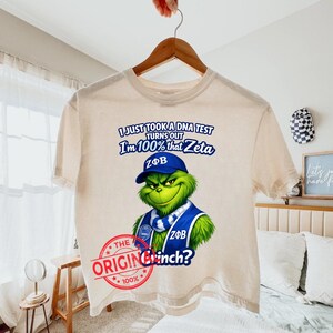 以下が含まれることがあります： クリーム色のTシャツ。青い帽子とベストを着た、Grinchのようなキャラクターが特徴です。「I'm 100% that Zeta」と「The Original Grinch?」の文字が赤いスタンプデザインでプリントされています。