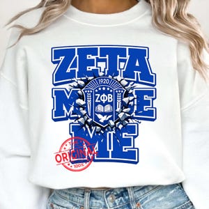 May include: White sweatshirt with a large blue graphic that reads "ZETA MIE" with a crest design in the center. The crest includes the text "1920 ZFB" and an open book. A red circular stamp that says "ORIGINAL 100%" is at the bottom.