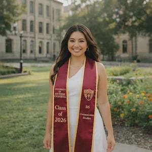 May include: A smiling person wearing a white dress and a maroon graduation sash with gold trim. The sash reads "University of Southern California," "Class of 2026," and "BS Communication Studies." The background includes a building and greenery.