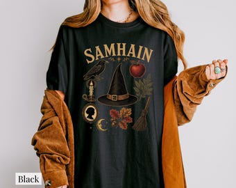 May include: A black t-shirt featuring the text SAMHAIN in gold lettering above a graphic illustration of a witch hat, a raven, a red apple, a lit candle, a cameo silhouette, a crescent moon, oak leaves, and a broomstick in earthy tones.