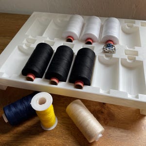 May include: A white plastic organiser tray holds spools of black and white thread. Additional spools of blue, yellow, and beige thread are scattered around the tray. A small metal bobbin is also present, suggesting a sewing or crafting theme.