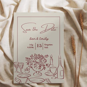 May include: A cream-coloured "Save the Date" card with burgundy script and illustrations of a table setting. The card includes the names "Ben & Emily", the date, time, and location. Two stalks of wheat are on the right.