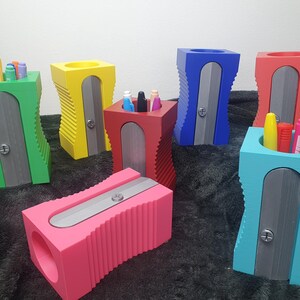 May include: A set of six colourful pencil sharpeners, each in a different colour: green, yellow, red, blue, pink and turquoise. The sharpeners are designed to look like traditional pencil sharpeners, with a metal blade and a hole for inserting pencils. The sharpeners are all arranged in a row on a grey surface.