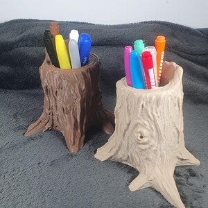 May include: Two pen holders shaped like tree stumps, one brown and one beige, filled with pens and markers. The holders have a textured, wood-like appearance, with detailed bark and root features. The pens include various colours like yellow, blue, and orange.