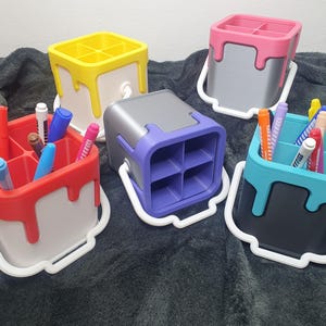 May include: Five paint can shaped desk organisers in different colours, each with a white handle and a drip effect. The organisers are filled with coloured markers.