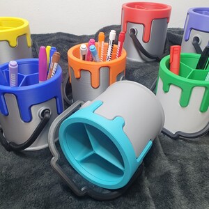 May include: Six paint can shaped desk organisers in different colours, each with a different colour of paint dripping down the side. The organisers have three compartments for pens, pencils, and other office supplies.