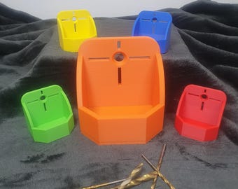 May include: Five colourful plastic drill bit holders, one orange, one yellow, one green, one red, and one blue. The holders are arranged in a semi-circle on a grey surface. There are five drill bits lying on the surface.