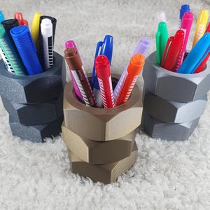 May include: Three hexagonal pen holders in grey, bronze, and silver, each filled with colourful pens and markers. The holders are stacked, resembling large nuts, and sit on a white, textured surface. The pens are various colours, including blue, red, green, and yellow.