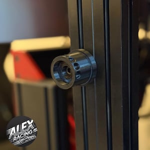 Op de afbeelding: Een close-up van een zwart metalen onderdeel met drie kleine gaten, bevestigd aan een zwart metalen frame. Het onderdeel is cilindrisch met een cirkelvormige opening. Het Alex Racing logo is zichtbaar linksonder.