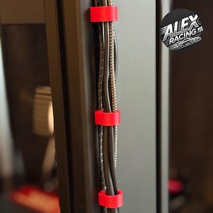 Könnte beinhalten: Nahaufnahme eines Computerturms mit schwarzen Kabeln, die mit drei roten Kabelclips befestigt sind. Die Kabel haben unterschiedliche Größen und Texturen. Ein Logo von "ALEX RACING" befindet sich in der oberen rechten Ecke.
