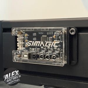 Puede incluir: Una placa de circuito SIMAGIC de acrílico transparente con componentes negros y texto blanco, incluyendo "SIMAGIC" y etiquetas de entrada. La placa está montada sobre una superficie negra. El logotipo de Alex Racing es visible en la esquina inferior izquierda.