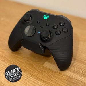Puede incluir: Un mando inalámbrico Xbox Elite Series 2 negro con agarres texturizados y un logotipo de Xbox verde. El mando tiene una placa frontal negra texturizada y un indicador luminoso azul.