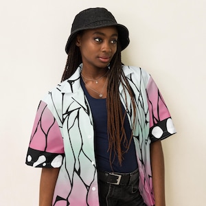 May include: A short-sleeved button-up shirt with a pink, white, and blue butterfly wing pattern. The shirt has a collar and a button closure. The model is wearing a black bucket hat and black jeans.