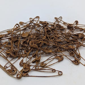 May include: A pile of rusty safety pins. The pins are all metal and have a brown, rusty color.