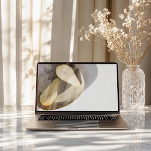 May include: A silver laptop sits on a marble table, displaying a photo of two pear halves on a plate. A clear, patterned glass vase holds dried flowers. Soft light filters through a sheer curtain in the background, creating a serene, minimalist aesthetic.