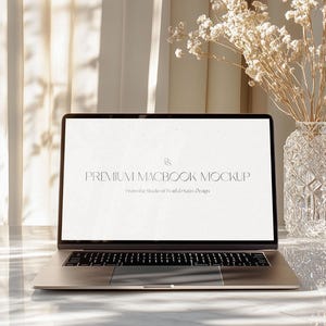 May include: A silver laptop with a lit screen displaying "PREMIUM MACBOOK MOCKUP" text. A clear glass vase with dried flowers sits to the right. The laptop is on a marble surface, with sunlight streaming through a sheer curtain.