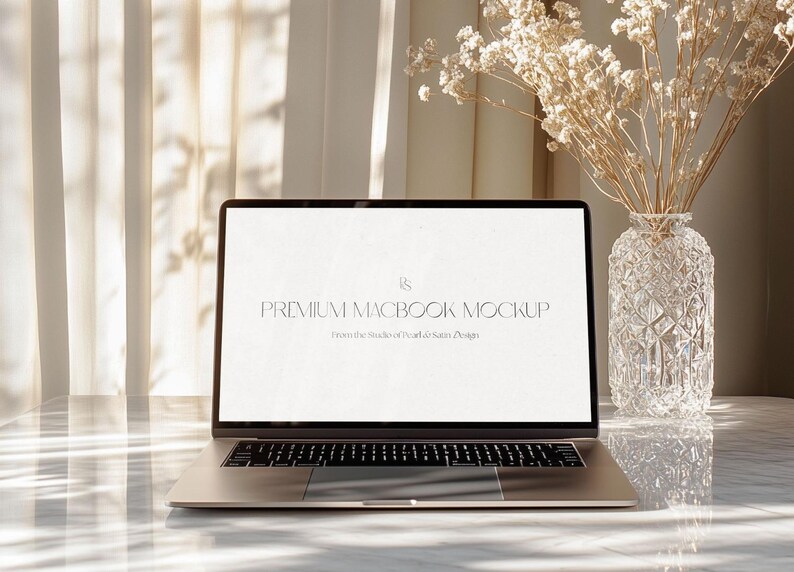 May include: A silver laptop with a lit screen displaying "PREMIUM MACBOOK MOCKUP" text. A clear glass vase with dried flowers sits to the right. The laptop is on a marble surface, with sunlight streaming through a sheer curtain.