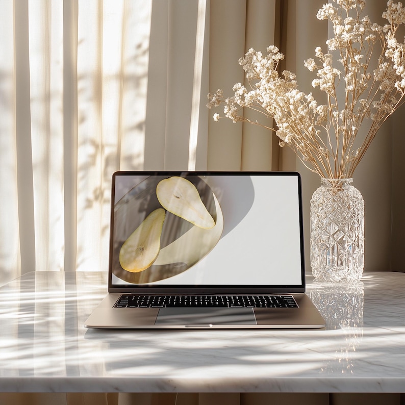 May include: A silver laptop sits on a marble table, displaying a photo of two pear halves on a plate. A clear, patterned glass vase holds dried flowers. Soft light filters through a sheer curtain in the background, creating a serene, minimalist aesthetic.