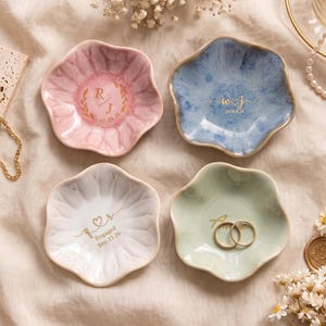 Peut inclure: Quatre coupelles à bijoux en céramique en forme de fleur, déclinées en rose pastel, bleu, blanc et vert clair. Chaque coupelle présente des inscriptions et symboles dorés personnalisés. L'une contient deux bagues dorées, toutes ont des bords ondulés.