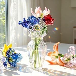May include: A clear glass vase filled with colourful glass flowers. The flowers include blue, red, pink, and yellow blooms. The vase is on a white surface near a window.