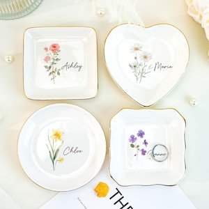 Puede incluir: Cuatro pequeños platos de cerámica blanca con bordes dorados. Cada plato presenta un diseño floral y un nombre: Ashley, Maria, Chloe y Anne. Los platos son cuadrados, en forma de corazón y redondos.