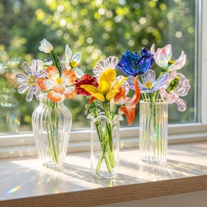 May include: Three glass vases containing colourful glass flowers. The flowers are in shades of orange, yellow, red, blue, and pink. The vases are on a wooden surface in front of a window.
