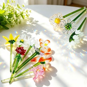May include: A selection of glass flower pens in various colours. The pens feature different flower designs, such as daisies, roses, and lilies, with green stems. The pens are displayed on a white surface, with sunlight creating colourful refractions.