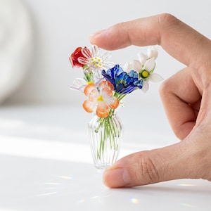 May include: A small glass vase holding a collection of colourful glass flowers. The arrangement features red, white, blue, and orange blooms. The clear vase has a ribbed design, displaying the delicate stems of the flowers.