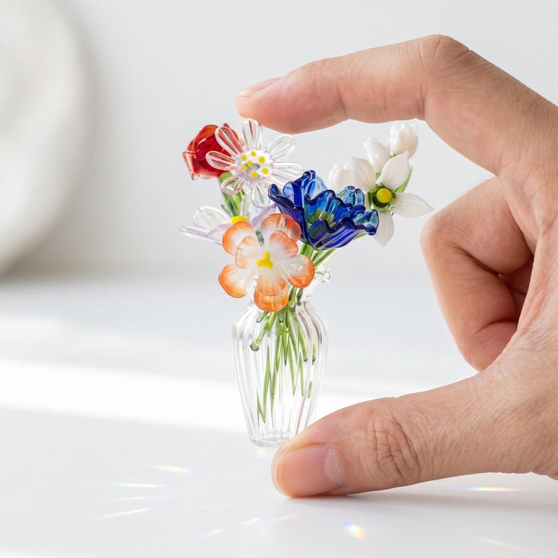 May include: A small glass vase holding a collection of colourful glass flowers. The arrangement features red, white, blue, and orange blooms. The clear vase has a ribbed design, displaying the delicate stems of the flowers.