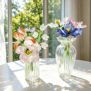 May include: Two glass vases on a white table, each holding colourful glass flowers. One vase has orange, white, and pink flowers. The other vase has blue, pink, and white flowers. The vases are clear with vertical lines.